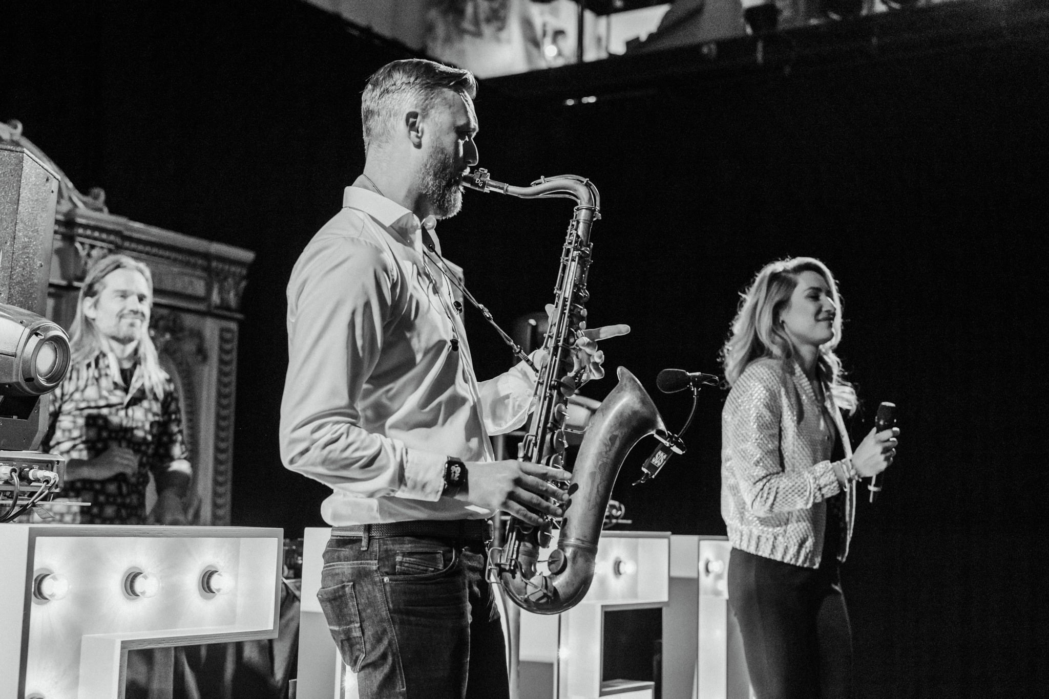 Een man speelt saxofoon terwijl een vrouw met een microfoon ernaast staat op een podium en de DJ achter grote LED letters.