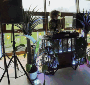 Filmpje van een DJ die draait achter een verlichte rotan DJ-booth met grote kamerplanten en speakers.